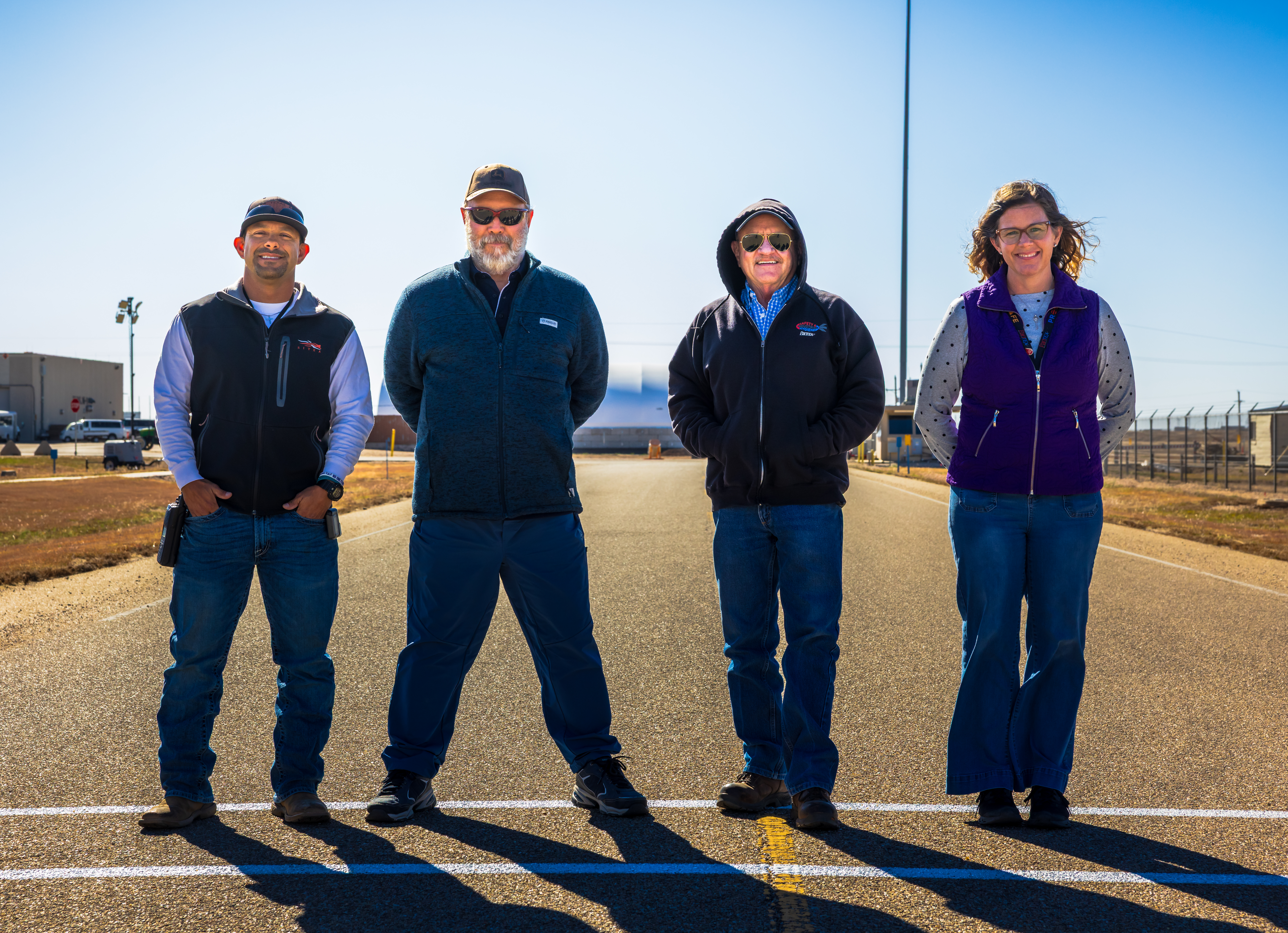 Three men and one woman stand in the middle of a roadway on a walking path, facing the camera and smiling. 