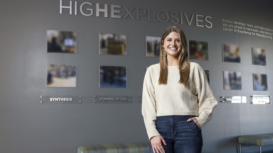 A young blonde woman stands in front of a grey wall lined with photographs. Above her, the words “high explosives” are visible in silver text.