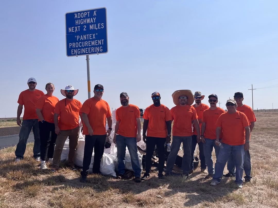 A group of men and women wearing bright orange T-shirts stand in front of a large pile of white trash bags under a road sign reading, “Adopt-A-Highway next two miles. Pantex Procurement Engineering.”